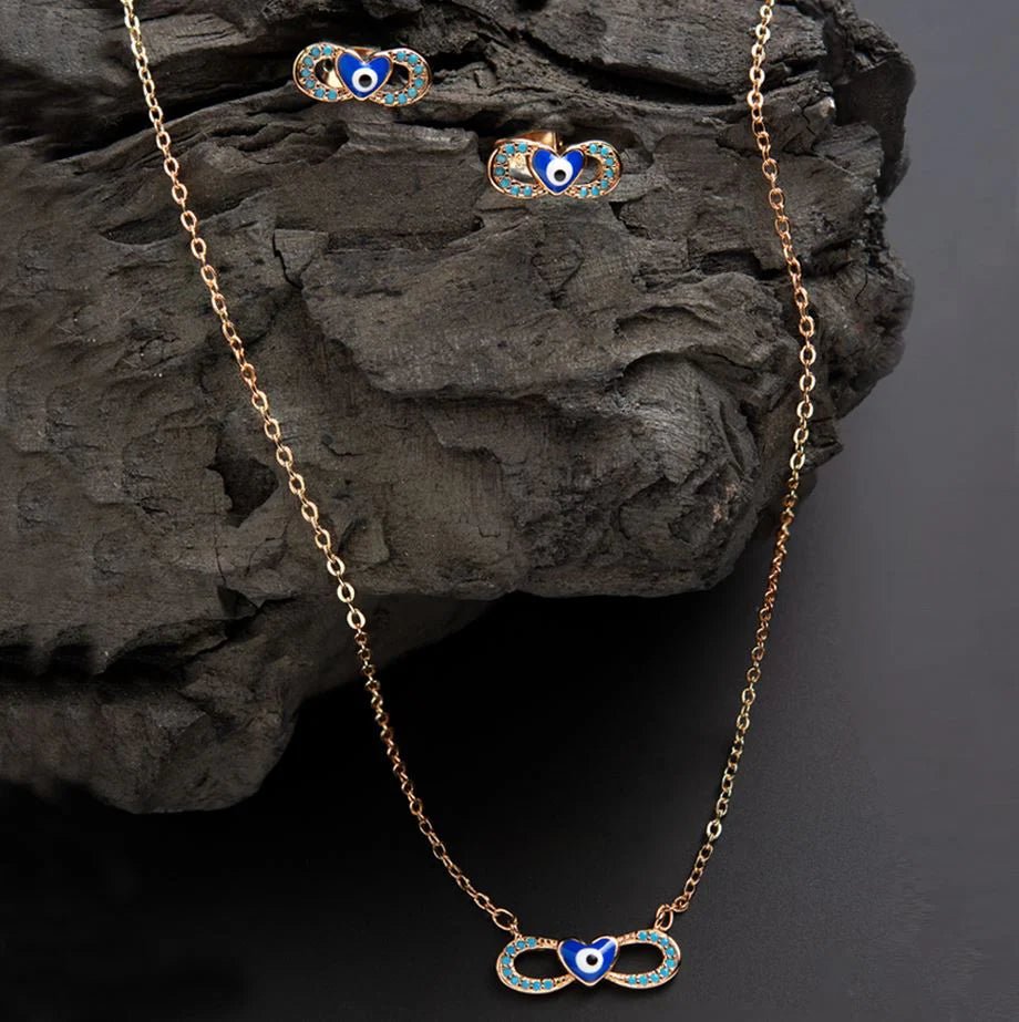 Gold-plated pendant set including earrings and a necklace, both featuring an infinity design with light blue enamel dots and a central heart in dark blue enamel with an eye detail.